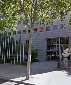 A modern office building with glass facade, surrounded by trees and parking spaces. - Olive Oil Times