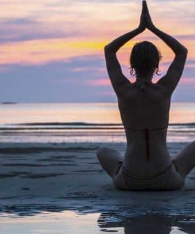 Individual sitting in a yoga pose on the beach during sunset with hands raised. - Olive Oil Times