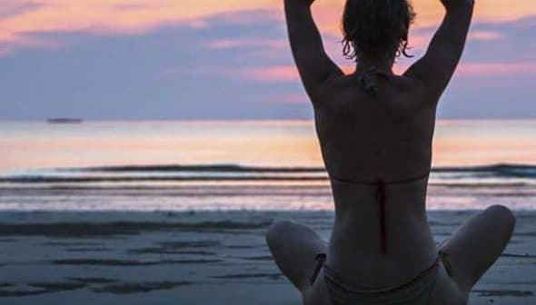 Individual sitting in a yoga pose on the beach during sunset with hands raised. - Olive Oil Times