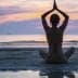 Individual sitting in a yoga pose on the beach during sunset with hands raised. - Olive Oil Times