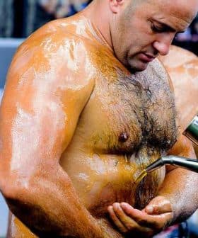 A man with oiled skin receiving oil poured onto his abdomen during a competition. - Olive Oil Times