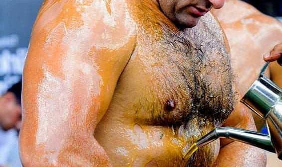A man with oiled skin receiving oil poured onto his abdomen during a competition. - Olive Oil Times