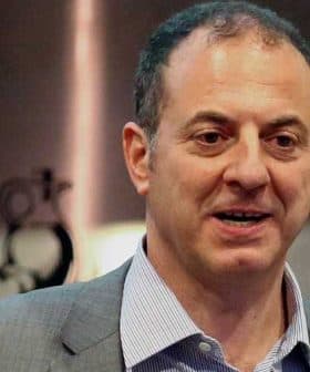 Man wearing a gray suit and striped shirt speaking at an event, with a neutral expression. - Olive Oil Times