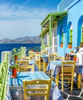 Outdoor dining area featuring tables and chairs in front of a blue and green building by the sea. - Olive Oil Times