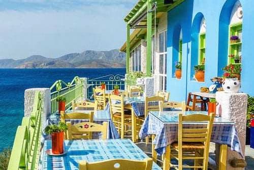 Outdoor dining area featuring tables and chairs in front of a blue and green building by the sea. - Olive Oil Times