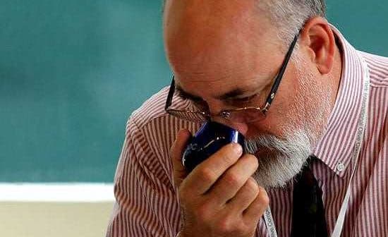 A man with glasses and a beard holding a blue object close to his face in a classroom setting. - Olive Oil Times