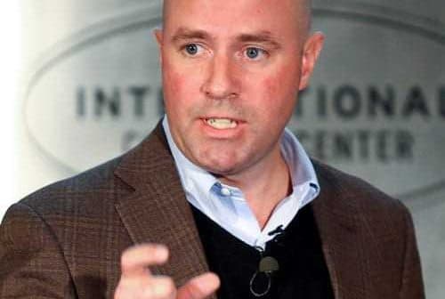 A man in a brown blazer and black sweater speaking at a podium during a conference. - Olive Oil Times