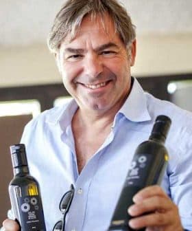 A man smiling while holding two bottles of olive oil in each hand. - Olive Oil Times