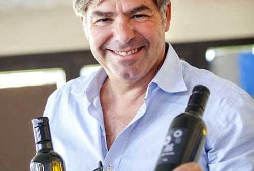 A man smiling while holding two bottles of olive oil in each hand. - Olive Oil Times