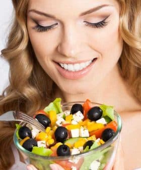 Woman with long blonde hair smiling while holding a bowl of colorful salad with vegetables and olives. - Olive Oil Times
