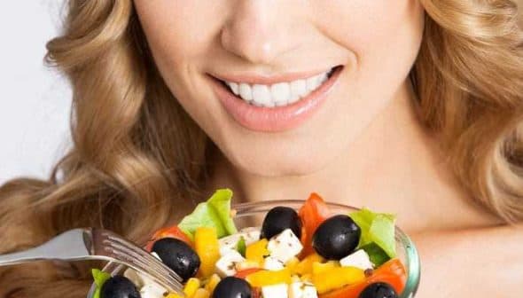 Woman with long blonde hair smiling while holding a bowl of colorful salad with vegetables and olives. - Olive Oil Times