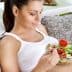 A woman sitting on a couch holding a bowl of salad with a fork, wearing a white tank top. - Olive Oil Times