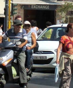 Two individuals on a scooter and a woman walking on a city street with vehicles in the background. - Olive Oil Times