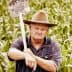 A man wearing a hat and overalls holding a shovel in a cornfield. - Olive Oil Times