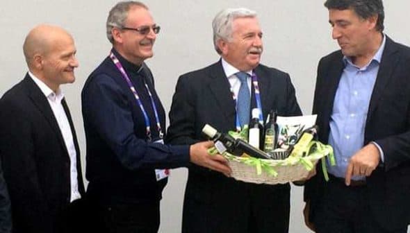 Four men standing together, one presenting a basket filled with olive oil products. - Olive Oil Times