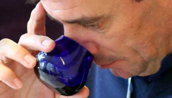 Man holding a blue glass cup and smelling the contents while tasting olive oil. - Olive Oil Times