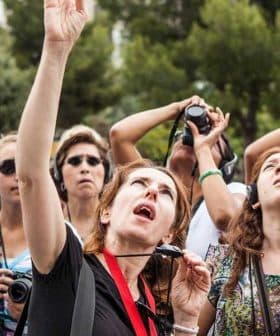 A group of tourists looking up at a landmark while wearing headphones and holding cameras. - Olive Oil Times