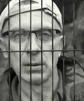 A man wearing glasses and a cap, looking through metal bars with a serious expression. - Olive Oil Times