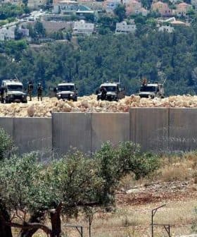Two military vehicles stationed near a concrete barrier with barbed wire in a hilly area. - Olive Oil Times