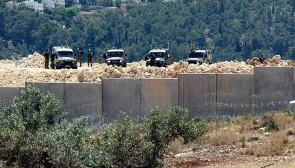 Two military vehicles stationed near a concrete barrier with barbed wire in a hilly area. - Olive Oil Times