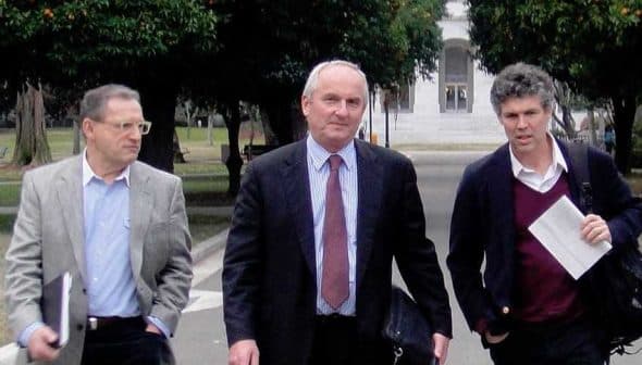 Three men walking together in a park, carrying briefcases and folders. - Olive Oil Times