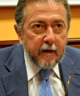 Man with gray hair and beard wearing a blue suit speaking at a conference table. - Olive Oil Times