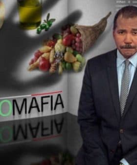 A man in a suit standing next to a graphic titled 'Agro Mafia' with food items and a glass of wine. - Olive Oil Times