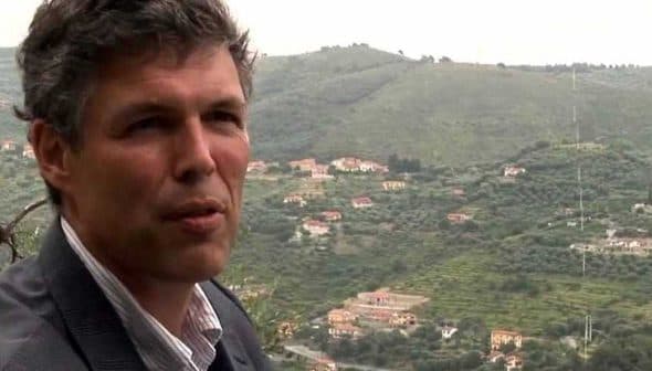 A man in a suit standing in front of an olive grove on a hillside. - Olive Oil Times