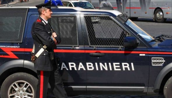A Carabinieri officer standing next to a marked police vehicle in an urban setting. - Olive Oil Times