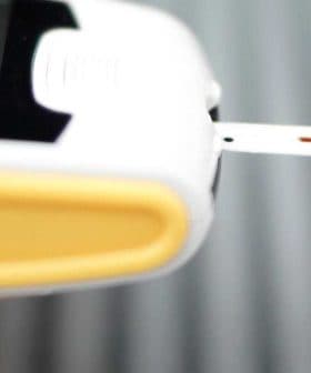 Person using a blood glucose meter with a test strip inserted and a drop of blood on their fingertip. - Olive Oil Times