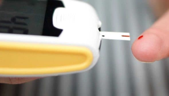 Person using a blood glucose meter with a test strip inserted and a drop of blood on their fingertip. - Olive Oil Times