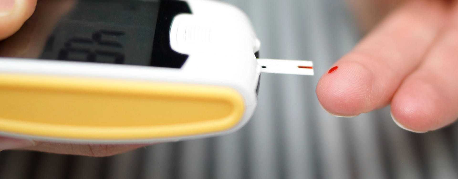 Person using a blood glucose meter with a test strip inserted and a drop of blood on their fingertip. - Olive Oil Times