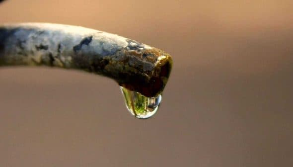 Close-up of a water droplet hanging from the tip of a faucet spout. - Olive Oil Times