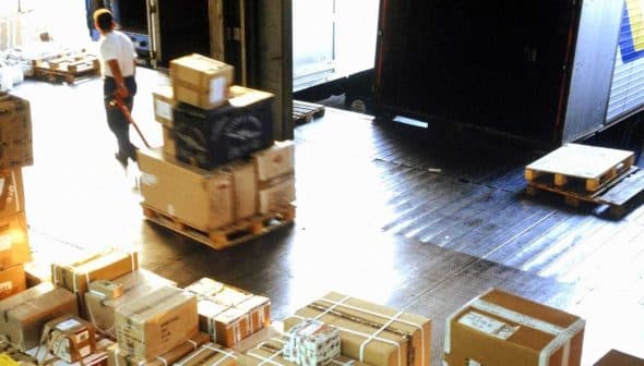 A warehouse interior with stacked packages and a worker using a pallet jack to move boxes. - Olive Oil Times