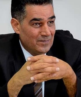 Man in a black suit sitting with hands clasped, smiling slightly while looking forward. - Olive Oil Times