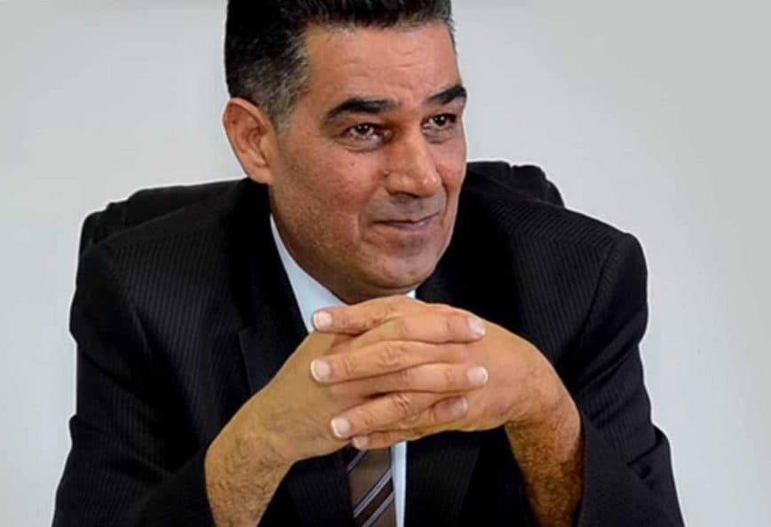 Man in a black suit sitting with hands clasped, smiling slightly while looking forward. - Olive Oil Times