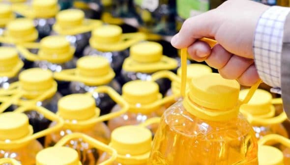 A hand gripping a bottle of oil with a yellow cap among multiple bottles on a shelf. - Olive Oil Times