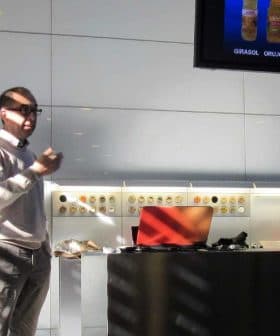 A person giving a presentation in a modern kitchen with a screen displaying various oil types. - Olive Oil Times