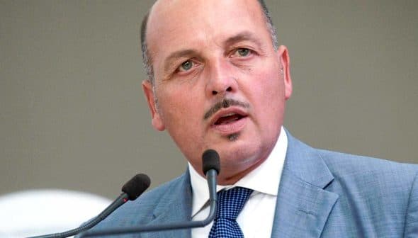 Man in a gray suit speaking into a microphone at a podium during an event. - Olive Oil Times