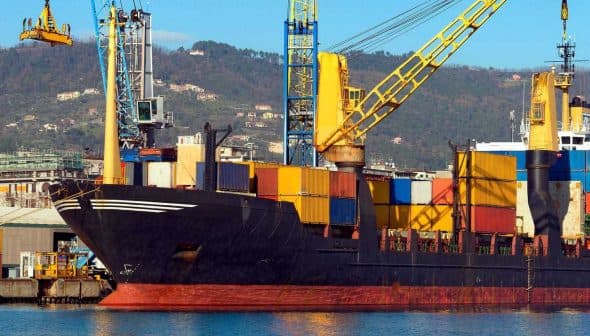 Cargo ship docked at a port with various colorful shipping containers stacked on board. - Olive Oil Times