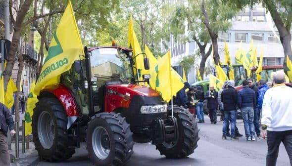 Red tractor surrounded by yellow flags at a demonstration in an urban area. - Olive Oil Times