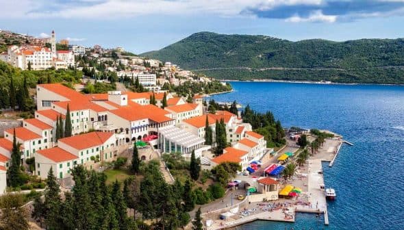 A coastal view of a Mediterranean town with red-roofed buildings and a blue sea. - Olive Oil Times