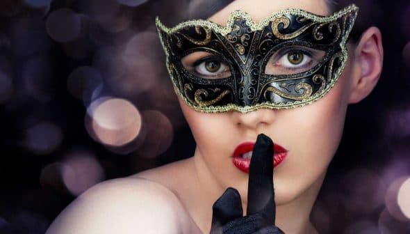 Woman with a black masquerade mask and finger on lips, signaling silence. - Olive Oil Times