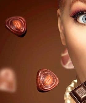 Woman with light skin holding a piece of chocolate with various chocolate shapes around her. - Olive Oil Times