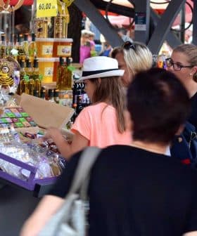 People shopping at a market with various products displayed on tables and vendors interacting with customers. - Olive Oil Times
