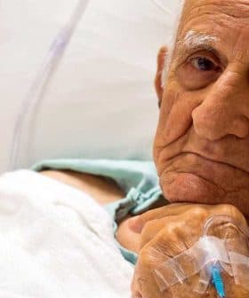 Elderly man lying in a hospital bed with an intravenous line in his hand. - Olive Oil Times