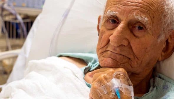 Elderly man lying in a hospital bed with an intravenous line in his hand. - Olive Oil Times