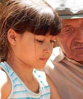A young girl and an elderly man sitting together, focused on an activity outdoors. - Olive Oil Times