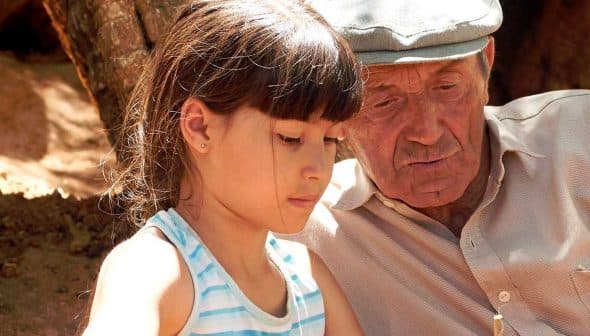 A young girl and an elderly man sitting together, focused on an activity outdoors. - Olive Oil Times
