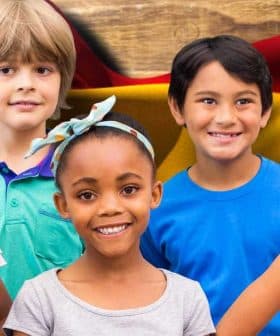Five children posing together in front of a Spanish flag background, smiling and wearing casual clothing. - Olive Oil Times